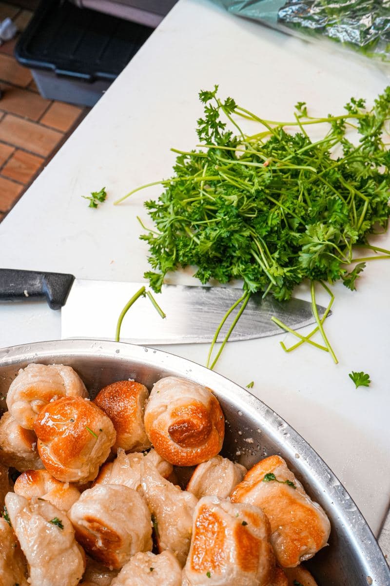 Garlic knots in a steel bowl beside fresh parsley on the cutting board